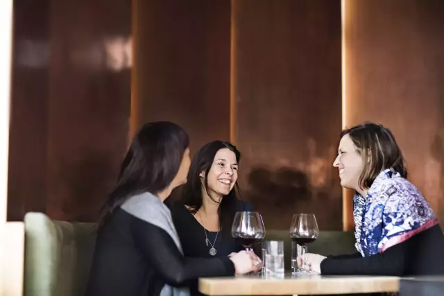 Drei Freundinnen sitzen gutgelaunt gemeinsam am Tisch einer Weinbar. Es wird Rotwein getrunken und gelacht.